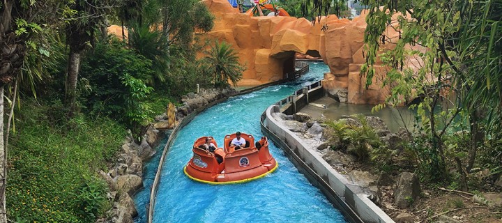 Foto de um bote redondo ocupado por 5 pessoas fluindo em um rio artificial na cor azul em direção à uma passagem coberta. Atração do Beto Carrero World, parque de diversões