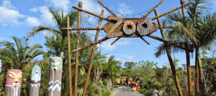 Foto da entrada de um zoológico com uma placa de madeira pendurada no arco de entrada com os dizeres “Zoo”